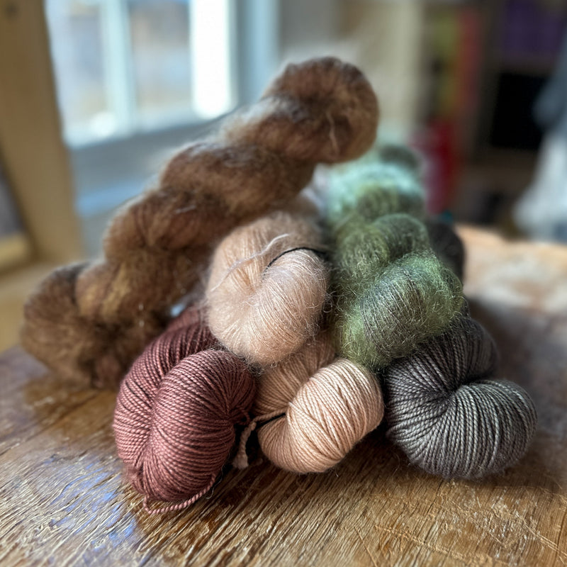 A soft palette of yarn skeins in three colors, arranged on a cool white marbled background with a sprig of greenery
