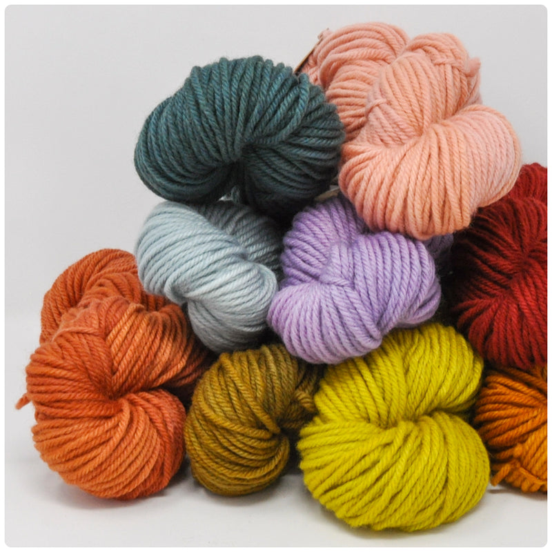 A soft palette of yarn skeins in three colors, arranged on a cool white marbled background with a sprig of greenery